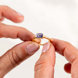 Light purple tanzanite ring in yellow gold, featuring an elegant east-west bezel setting, held between fingers.