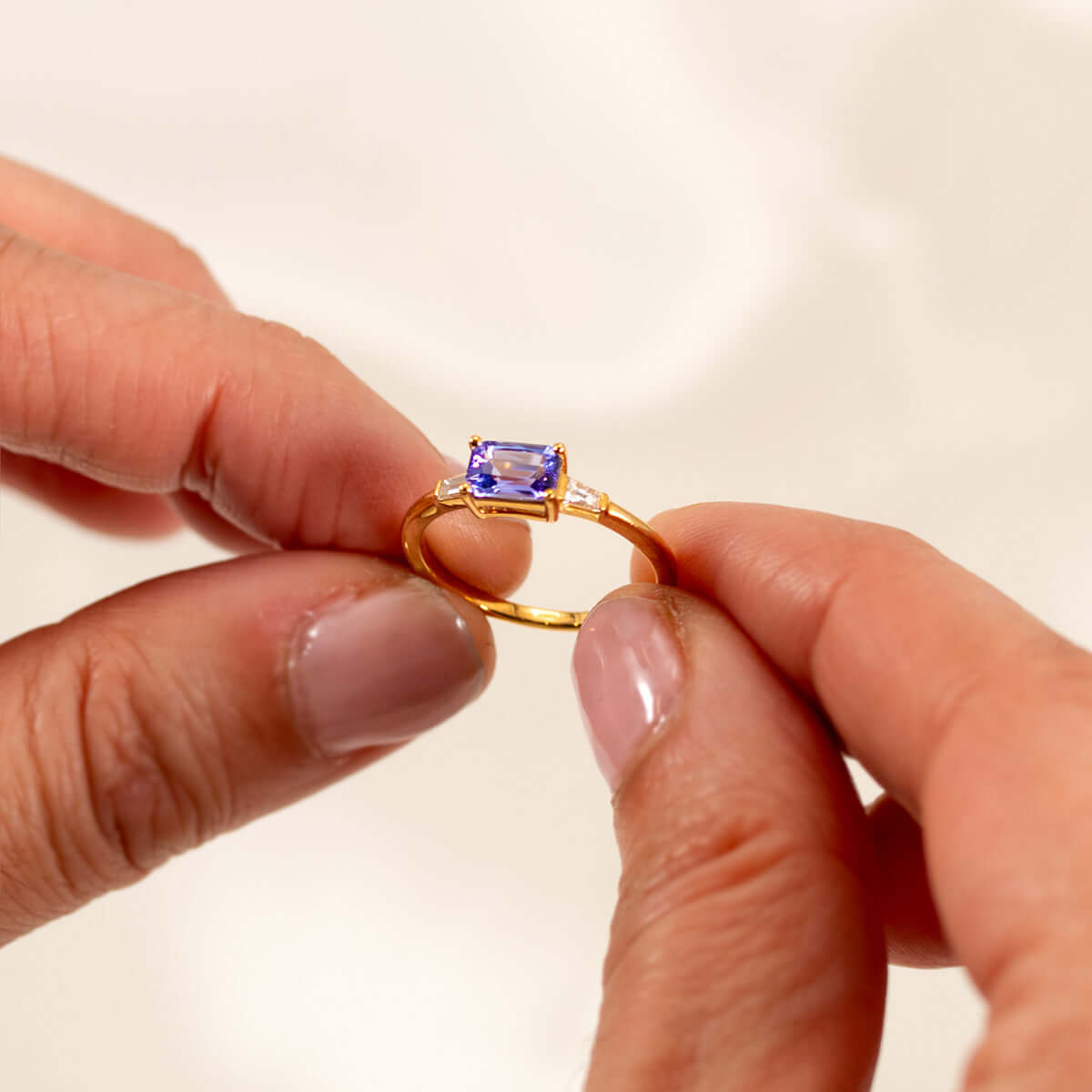 Elegant light blueish-purple tanzanite east-west ring held by hands, set in 14K yellow gold with baguette diamonds.